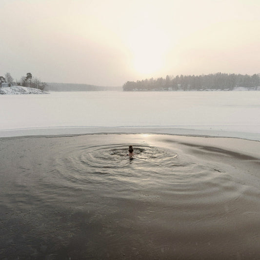 Person swimming in a hole cut in a frozen lake, snowy wooded shore beyond