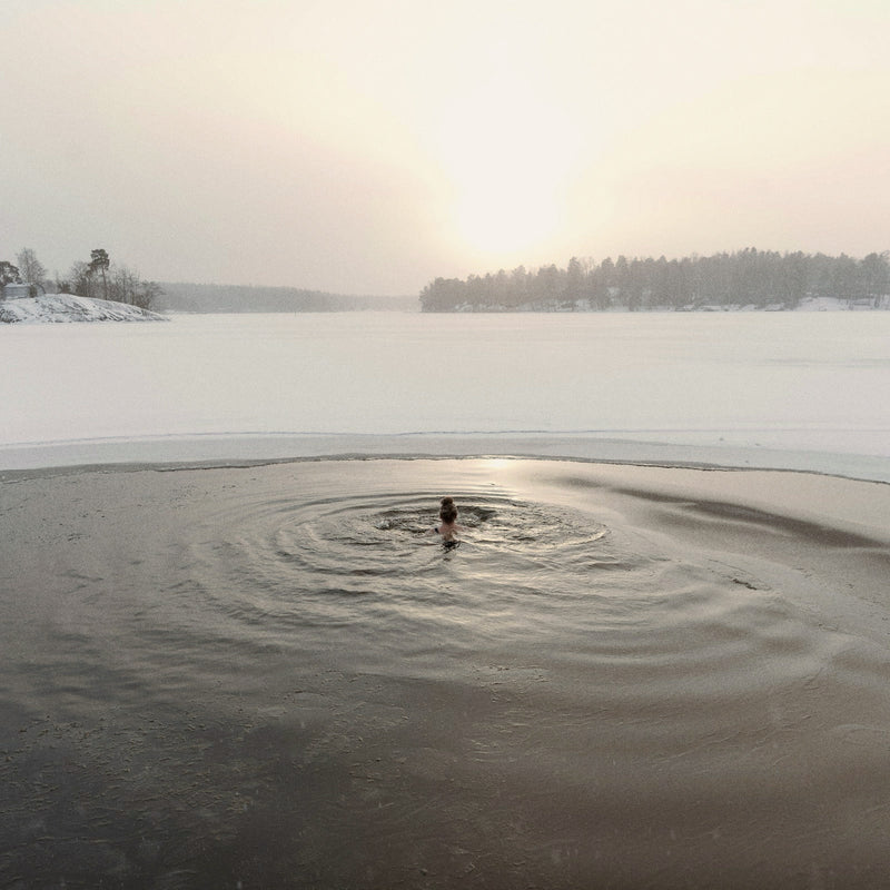 Person swimming in a hole cut in a frozen lake, snowy wooded shore beyond